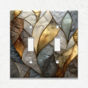 May include: A decorative double light switch cover with a mosaic design. The cover features a pattern of overlapping leaf-like shapes in shades of gold, brown, gray, and white, outlined with gold trim. The switch openings are rectangular.