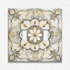 May include: Decorative double light switch cover with a floral design. The cover features a cream, gray, and gold color palette. The design includes a central flower and ornate, symmetrical patterns.