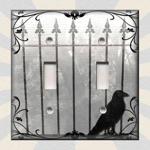 May include: A black and white light switch cover with a silhouette of a crow perched on a wrought iron gate. The gate has a gothic design with pointed tips and floral accents.