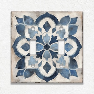 May include: Decorative double light switch cover featuring a blue and white floral design. The design is centered on a square plate with a neutral background. The floral pattern is symmetrical, with a central flower and surrounding petal shapes.