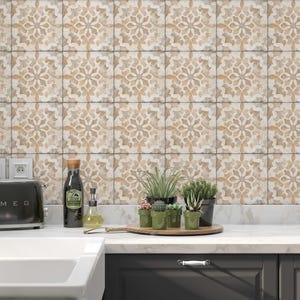 May include: A kitchen with white countertops and gray cabinets. The backsplash is made of beige and brown patterned tiles. There is a stainless steel range hood, a toaster, a sink, and a faucet. There are also plants in pots on the counter.