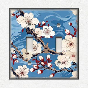 May include: A light blue double light switch cover with a floral design. The design features white cherry blossoms with red accents on a blue background.