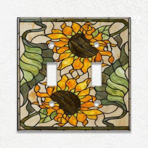 May include: A decorative double light switch cover featuring a stained glass design with two sunflowers. The sunflowers have yellow and orange petals with brown centers. Green leaves and stems surround the flowers, set against a cream-colored background.