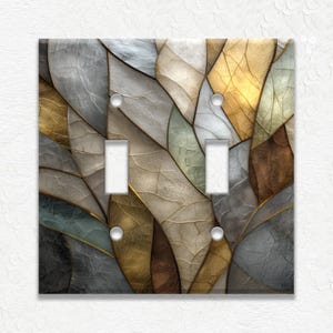 May include: A decorative double light switch cover with a leaf-like design. The cover features a mosaic of muted colors including gold, gray, brown, and green. The design has a textured appearance with gold-colored outlines.