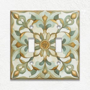 May include: A decorative double light switch cover with a floral design. The cover features a cream background with teal, gold, and olive green accents. The design is symmetrical, with a central flower and ornate leaf patterns.