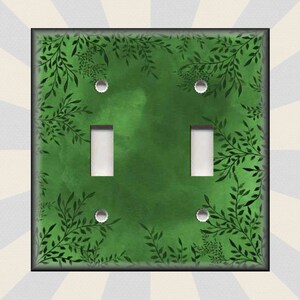 May include: A green light switch cover with a black leafy pattern. The switch cover has two toggle switches and two screw holes.