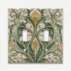May include: Decorative double light switch cover with an Art Nouveau design. The cover features a cream-colored background with a raised pattern of green and gold floral elements. The switch openings are white.