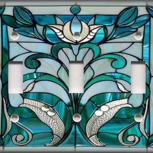 May include: A decorative light switch cover with a stained glass design. The design features teal, white, and silver floral and leaf patterns. The cover has three light switches and a dark border.