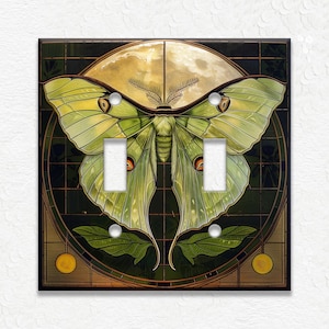 May include: A decorative double light switch cover featuring a large green moth with yellow accents, set against a backdrop of a full moon and dark green grid. The design includes leaves and circular elements.
