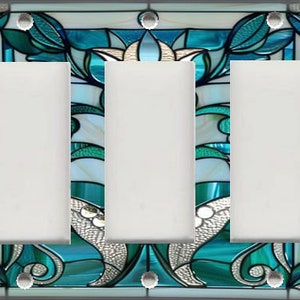 May include: A decorative triple light switch cover with a stained glass design. The design features teal, white, and silver floral and abstract patterns. The cover has three blank white switch openings and silver screw heads.