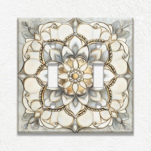 May include: A decorative light switch cover featuring a floral design with a gold, white, and gray color palette. The cover has two toggle switch openings and a raised, textured surface. The design is symmetrical and detailed.