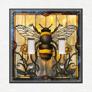 May include: A decorative light switch cover featuring a stained-glass design with a large bee in the center. The bee has yellow and brown stripes, with detailed wings. Yellow flowers and green leaves surround the bee, set against a yellow and blue background.