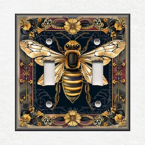 May include: A decorative light switch cover featuring a detailed illustration of a bee with gold and black stripes, surrounded by ornate floral designs in gold and red on a dark blue background.
