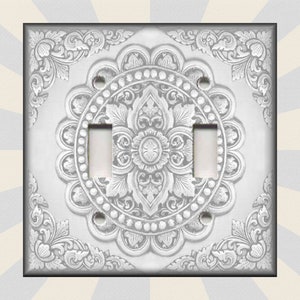 May include: A white light switch cover with a silver, ornate, floral design. The design features a central mandala with two toggle switches.