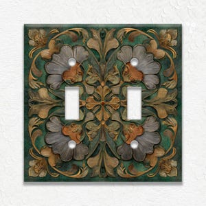 May include: A decorative double light switch cover with a floral and leaf design. The cover features a teal background with raised, detailed carvings in shades of gold, brown, and gray. The design includes two toggle switch openings.