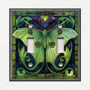 May include: A light switch cover featuring a stained glass design of a green luna moth with intricate details. The moth is surrounded by a dark blue background with floral accents.