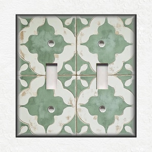 May include: A decorative double light switch cover featuring a distressed, floral tile design. The cover has a square shape with a black border. The tile pattern is in shades of green, white, and gold.