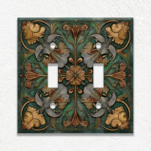 May include: A decorative double light switch cover with an ornate floral design. The cover features a square shape with a dark teal background and detailed gold, silver, and brown floral patterns. The switch openings are white.