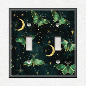 May include: A decorative light switch cover featuring a dark teal background with a celestial design. The cover is adorned with green and gold moths, crescent moons, and stars. The cover has two light switch openings.