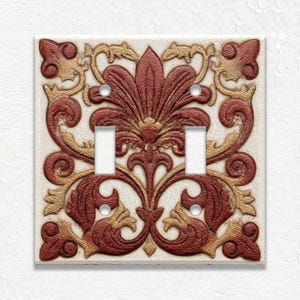 May include: A decorative double light switch cover with a detailed floral design. The cover features a cream-colored background with a raised pattern in shades of burgundy and gold. The design includes stylized leaves and symmetrical elements.