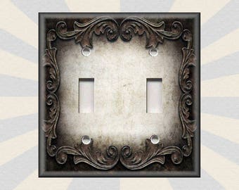 May include: A decorative light switch cover with a distressed, aged look. The cover features an ornate, intricate design with a dark brown finish. The switch plate has two toggle switches.