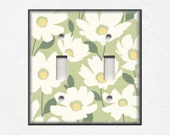 Puede incluir: Una cubierta de interruptor de luz floral verde claro y blanco con un fondo blanco y un patrón de flores blancas con centros amarillos. La cubierta del interruptor tiene dos interruptores de palanca.