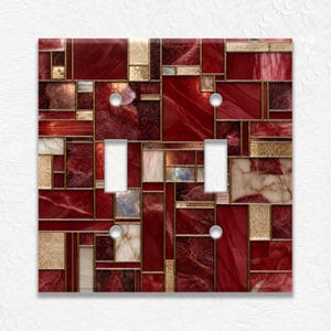 May include: A double light switch cover plate featuring a geometric mosaic pattern of deep red, burgundy, and white marble-like segments separated by thin gold lines, with two rectangular cutouts and four circular screw holes for installation.