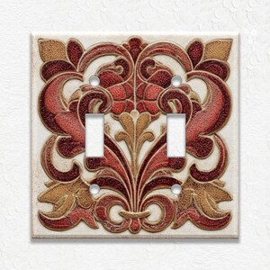 May include: A decorative double light switch cover with a raised floral design. The cover features a cream-colored background with a detailed pattern in shades of red, brown, and gold. The switch openings are white.