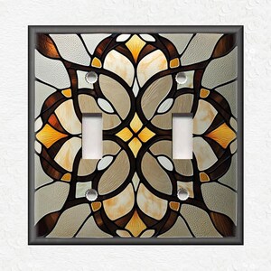 May include: A decorative double light switch cover with a stained glass design. The cover features a symmetrical floral pattern in shades of brown, beige, and yellow, framed by a dark gray border. The light switches are white.