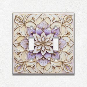 May include: A decorative light switch cover featuring a floral mandala design. The cover has a square shape with a cream-colored background and a detailed pattern in shades of purple, gold, and cream. It has two switch openings and a silver frame.