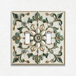 May include: Decorative double light switch cover with a floral design. The cover features a cream-colored background with raised floral patterns in shades of green and gold. The switch plate is square and has a gold-colored border.