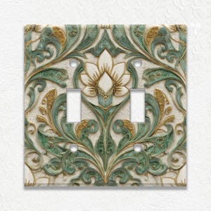 May include: A decorative double light switch cover with a floral design. The cover features a cream background with green and gold accents. The design includes a central flower and ornate scrollwork.