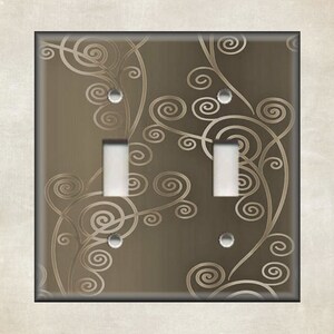 May include: A decorative double light switch cover with a brown background and a swirling, scroll-like design in a lighter beige color. The cover has two toggle switches and a neutral border.