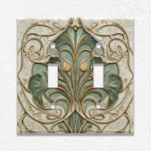 May include: Decorative double light switch cover with an Art Nouveau design. The cover features a cream-colored background with a raised, ornate pattern in gold and green. The design includes floral and leaf motifs.