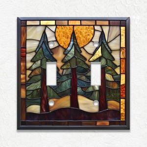 May include: A stained glass double light switch cover featuring a forest scene. The design includes three evergreen trees, a setting sun, and a dark brown frame. The trees are green and blue, with red trunks. The sun is yellow and orange.