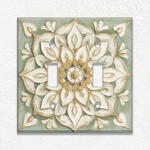 May include: A decorative double light switch cover with a floral design. The cover features a raised, cream-colored floral pattern with gold accents on a pale green background. The switch plate has two light switches and silver screws.