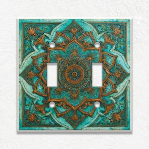 May include: Decorative double light switch cover with a teal and bronze mandala design. The cover features a detailed floral pattern with a central circular motif and two toggle switch openings. The design is set against a white background.