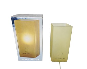 May include: A rectangular, off-white table lamp with a square base and a slightly angled design. The lamp is next to its original box, which features the lamp lit up. The text "JAN DES BOUVRIE" is printed on the lamp and box.