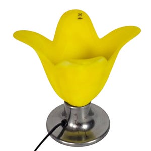 Op de afbeelding: Een gele glazen lamp in de vorm van een tulp met een geborstelde metalen voet. De lamp heeft een brede, open bovenkant en een gebogen, bloembladachtig ontwerp. Een zwart netsnoer loopt vanaf de voet. Decoratieve verlichting.
