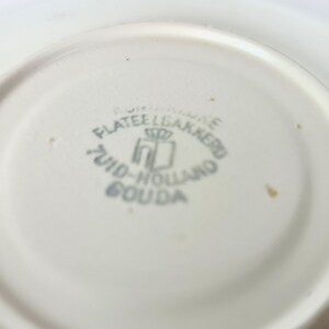 Pode incluir: Close-up da parte inferior de um prato de cer&acirc;mica branco com um logotipo azul e branco que diz "Plateelbakkerij Zuid-Holland Gouda".