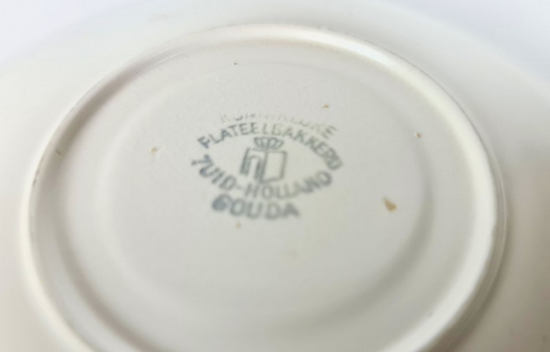 Pode incluir: Close-up da parte inferior de um prato de cer&acirc;mica branco com um logotipo azul e branco que diz "Plateelbakkerij Zuid-Holland Gouda".