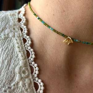 Può includere: Delicata collana di perline con perline verdi e turchese. Un piccolo ciondolo a forma di fiore dorato è appeso al centro. La collana è indossata su un top di pizzo bianco.