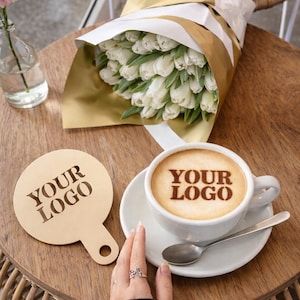 May include: A bouquet of white tulips wrapped in gold and white paper, a cup of coffee with "YOUR LOGO" in the foam, and a wooden coaster with "YOUR LOGO" on a wooden table. A small pink flower in a glass vase is also present.