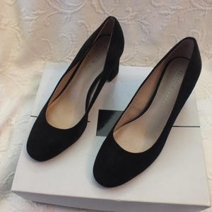 May include: A pair of black suede pumps with a low block heel. The shoes are on a white box.