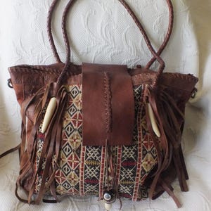 May include: Brown leather tote bag with a woven fabric panel featuring geometric patterns in red, black, and beige. The bag has long braided handles, leather fringe, and decorative bone-like accents.