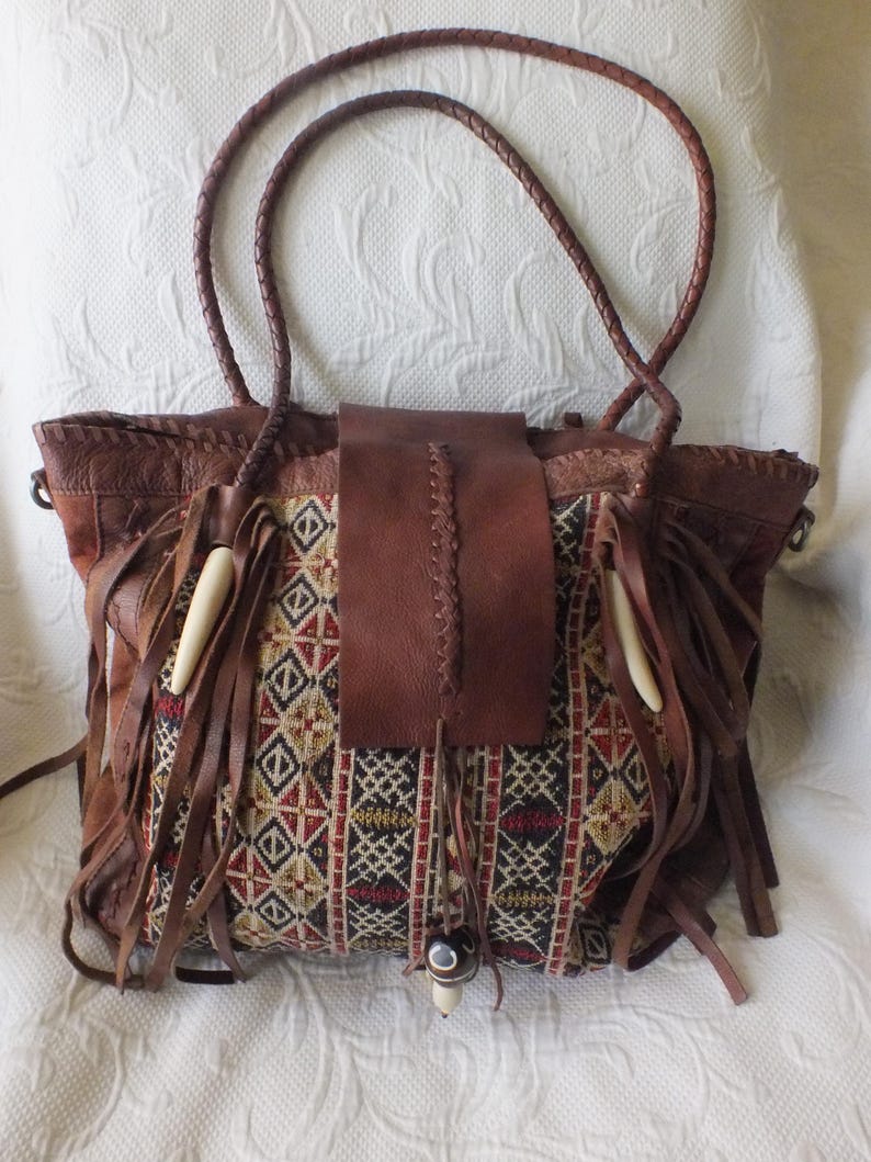 May include: Brown leather tote bag with a woven fabric panel featuring geometric patterns in red, black, and beige. The bag has long braided handles, leather fringe, and decorative bone-like accents.