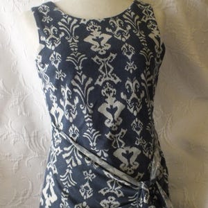May include: Sleeveless navy blue dress with a white, ornate pattern. The dress features a round neckline and a wrap-style design that ties at the waist. The fabric has a soft, flowing appearance, suitable for casual or semi-formal wear.