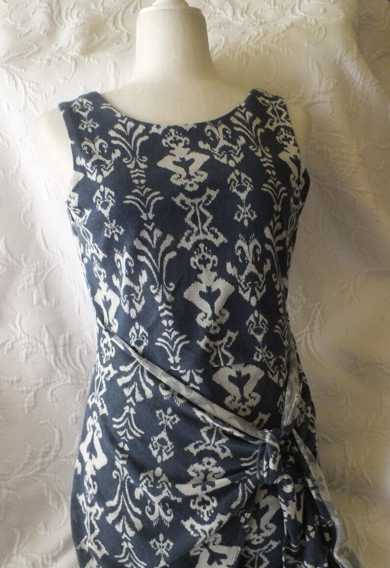 May include: Sleeveless navy blue dress with a white, ornate pattern. The dress features a round neckline and a wrap-style design that ties at the waist. The fabric has a soft, flowing appearance, suitable for casual or semi-formal wear.