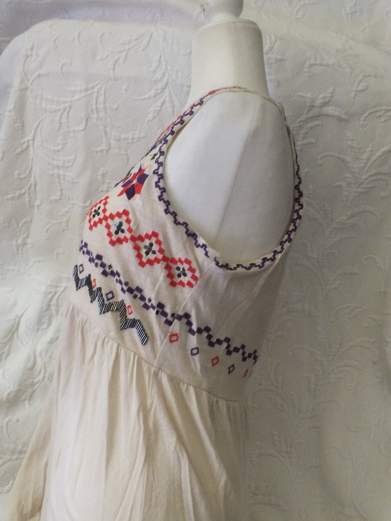 May include: A white sleeveless dress with a geometric pattern embroidered on the bodice in red, blue, and purple.