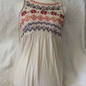 May include: A sleeveless, cream-colored, cotton dress with a geometric pattern embroidered on the neckline in shades of red, orange, purple, and black.